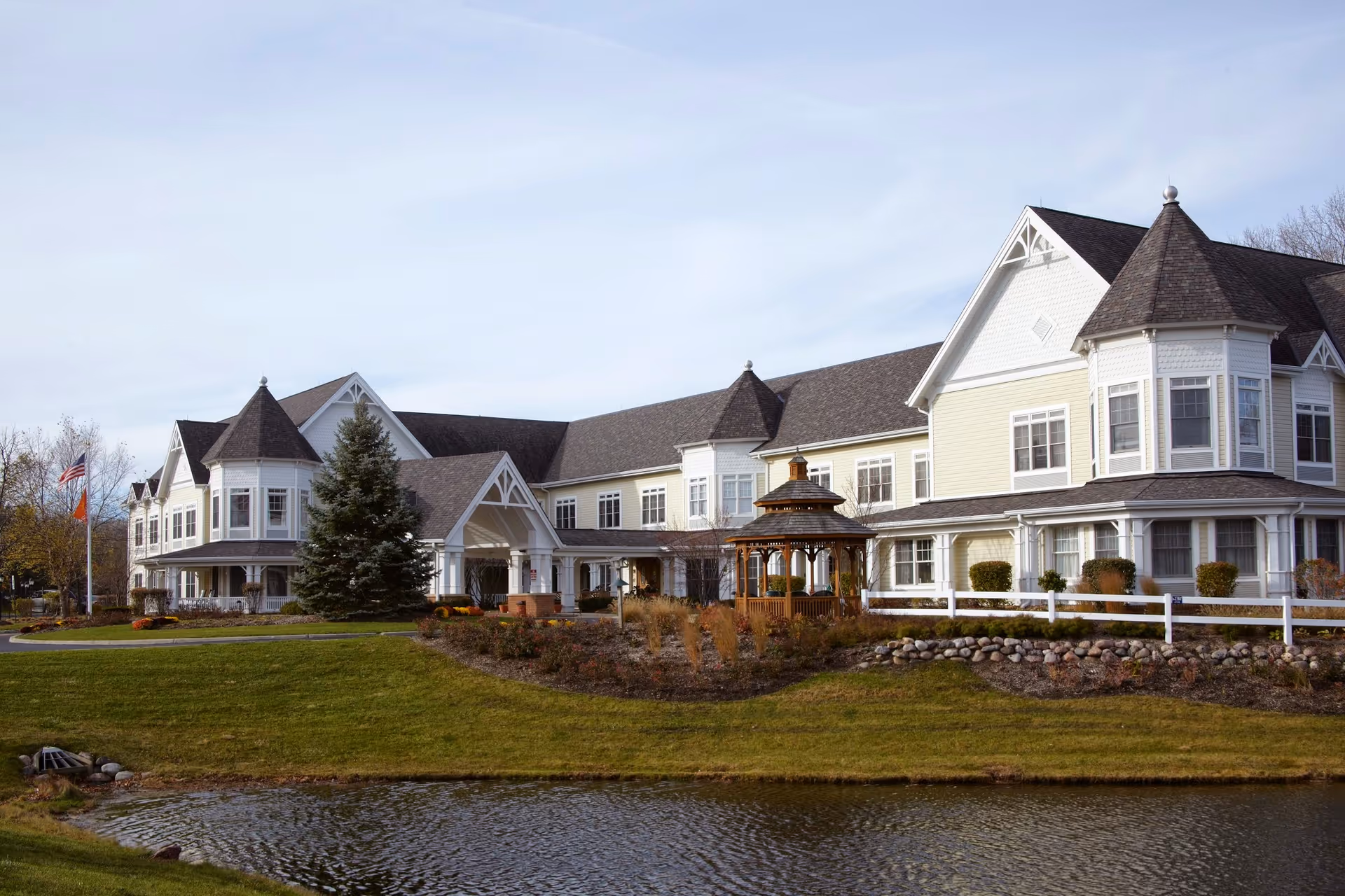 Exterior view of a large, elegant senior living facility building with white and beige siding, multiple gables, and a turret. The building is surrounded by well-maintained landscaping, including a small pond in the foreground, a wooden gazebo, and a white fence. Two flagpoles with flags are visible on the left side of the image under a partly cloudy sky.