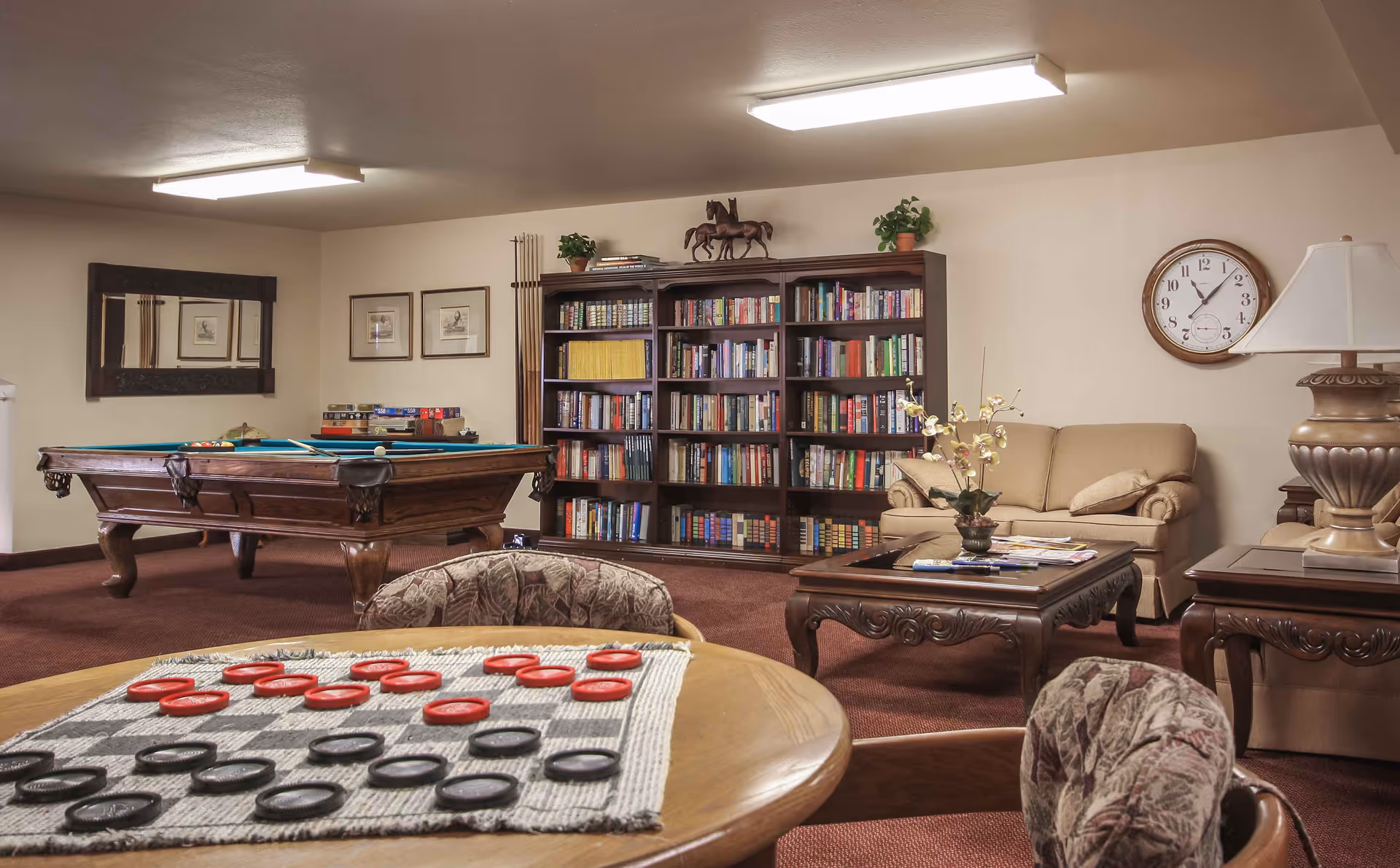 Community lounge featuring a pool table, large bookshelf, sofa and coffee table, with a table and checkers in the foreground.