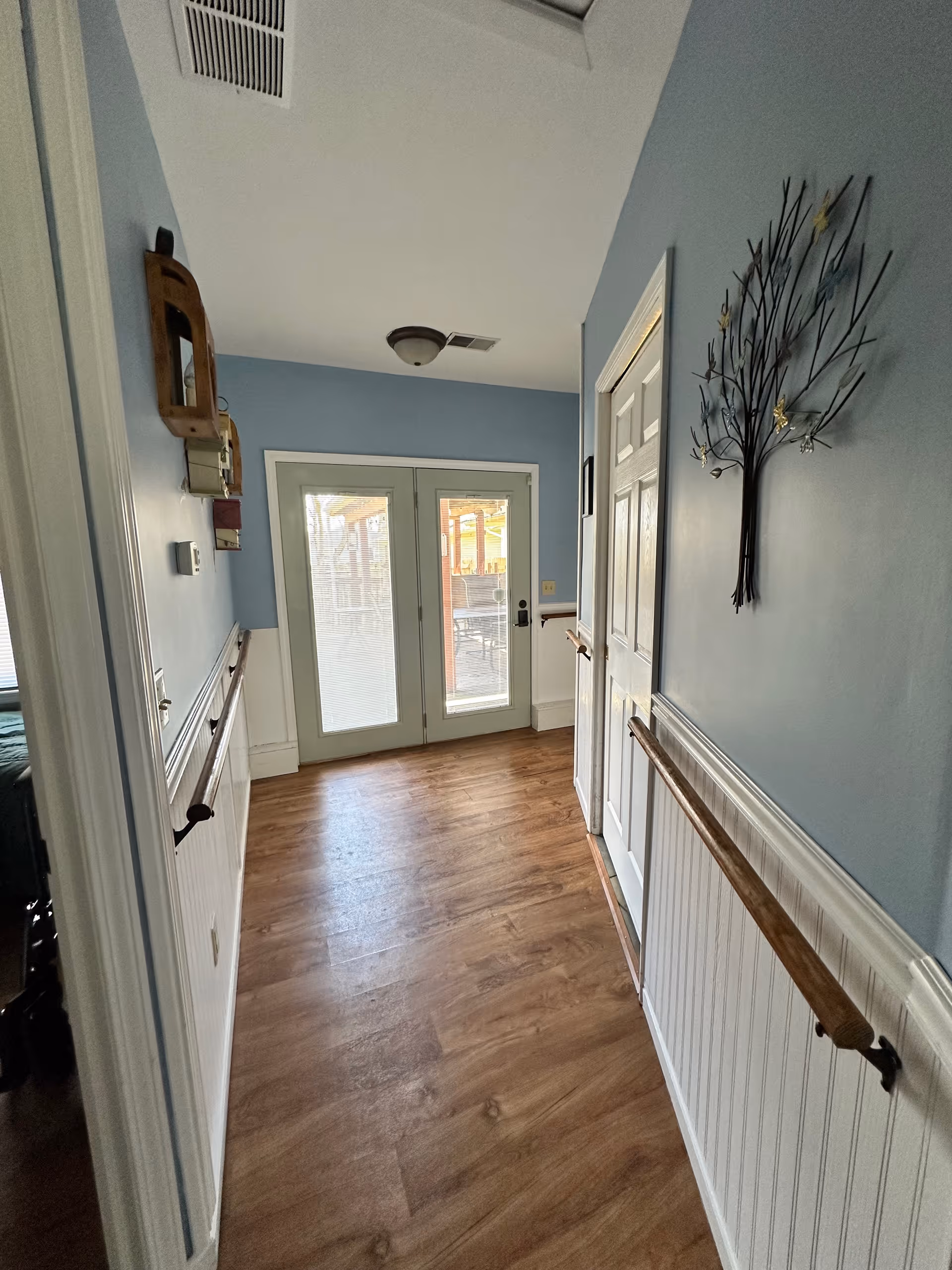 A hallway with light blue walls and wooden handrails on both sides. The floor is wood-patterned, and there is a decorative metal wall art shaped like a tree on the right wall. At the end of the hallway, there are double glass doors leading to an outdoor area with a patio and furniture visible through the glass.