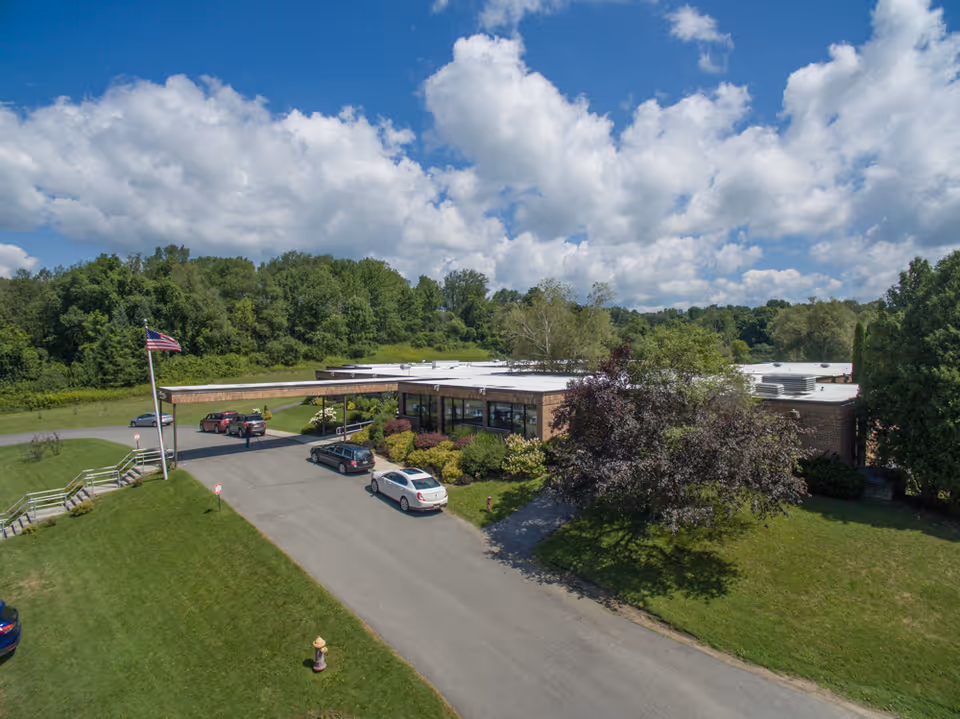 Aerial view of Alpine Rehabilitation & Nursing Center showing a single-story building with a flat roof surrounded by green lawns, trees, and a driveway with several parked cars. An American flag is flying on a flagpole near the entrance. The sky is partly cloudy with blue patches.