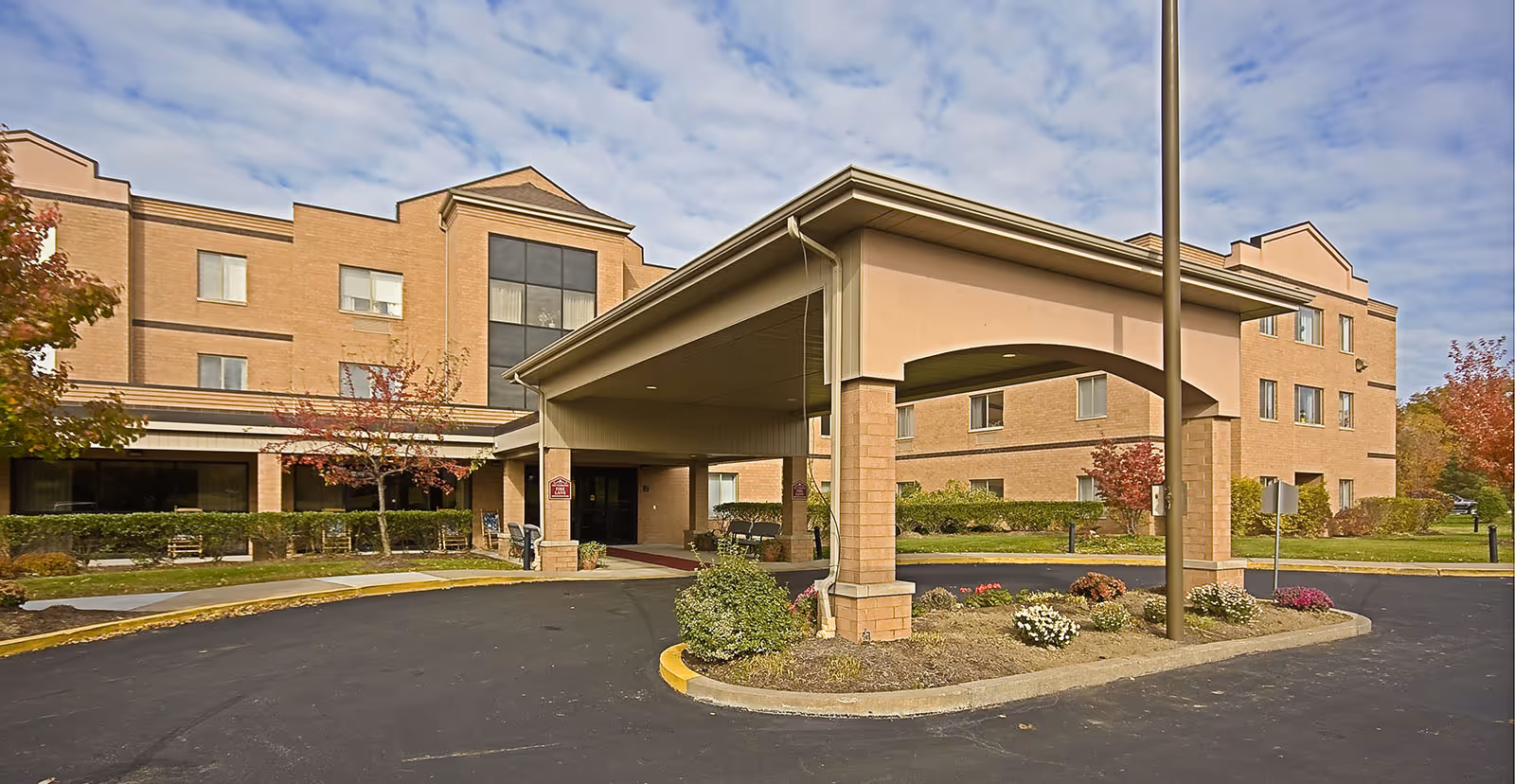 Exterior view of Holiday Niagara Village senior living facility showing a three-story brick building with a covered entrance driveway, landscaped flower beds, and trees with autumn foliage under a partly cloudy sky.