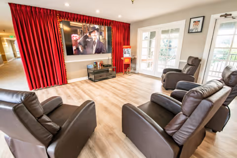 A bright and spacious living room area with four dark brown leather recliners arranged facing a large flat-screen TV mounted on the wall. The TV is framed by red curtains. There is a small black TV stand below the screen with some items on it, and a popcorn machine is placed near a set of glass doors leading outside. The room has light wood flooring and neutral-colored walls.