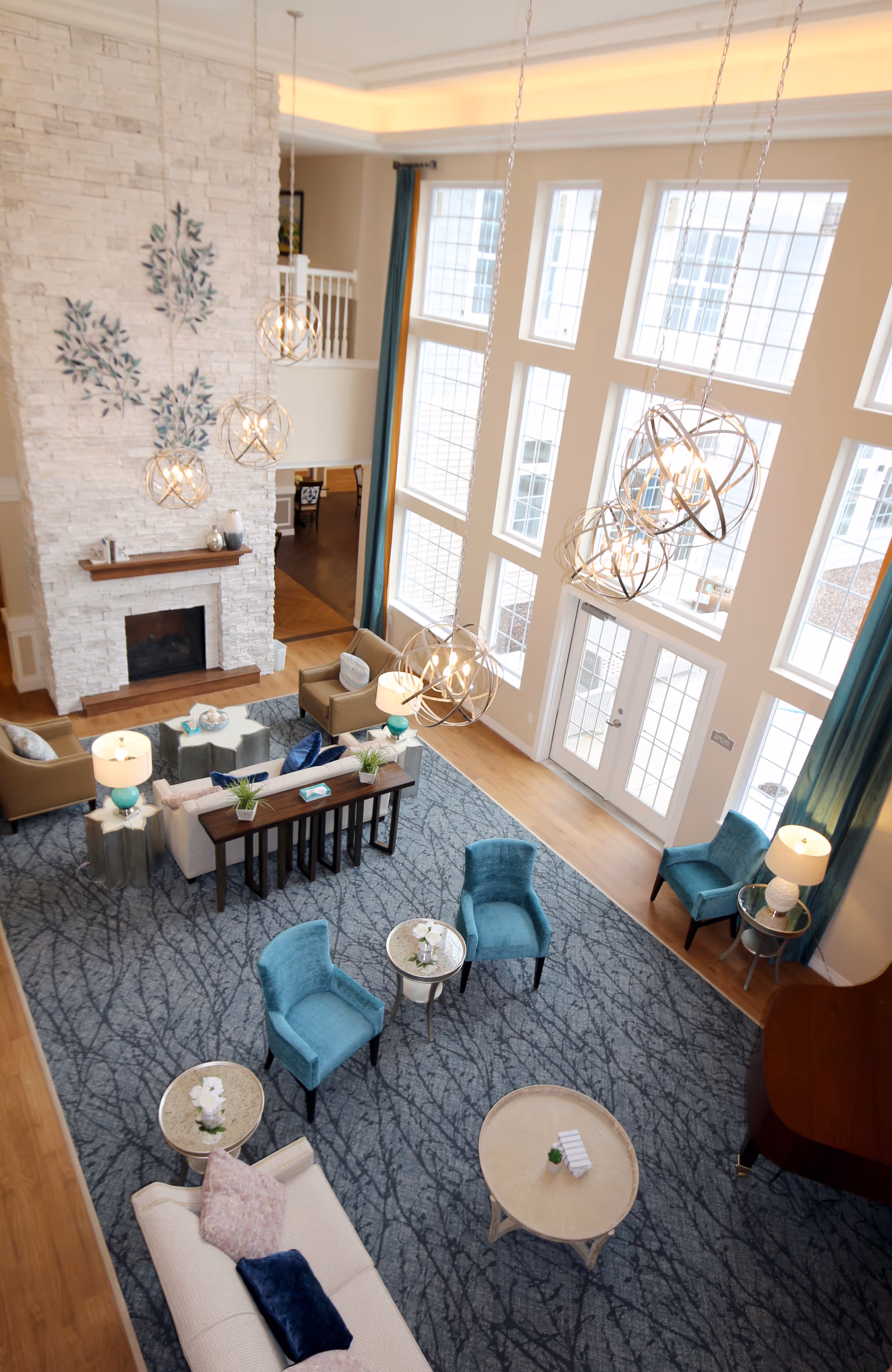 A spacious and well-lit living room area with large floor-to-ceiling windows, modern hanging light fixtures, a stone fireplace, multiple seating options including beige sofas and teal armchairs, small round tables with decorative plants, and a blue patterned carpet covering the floor.