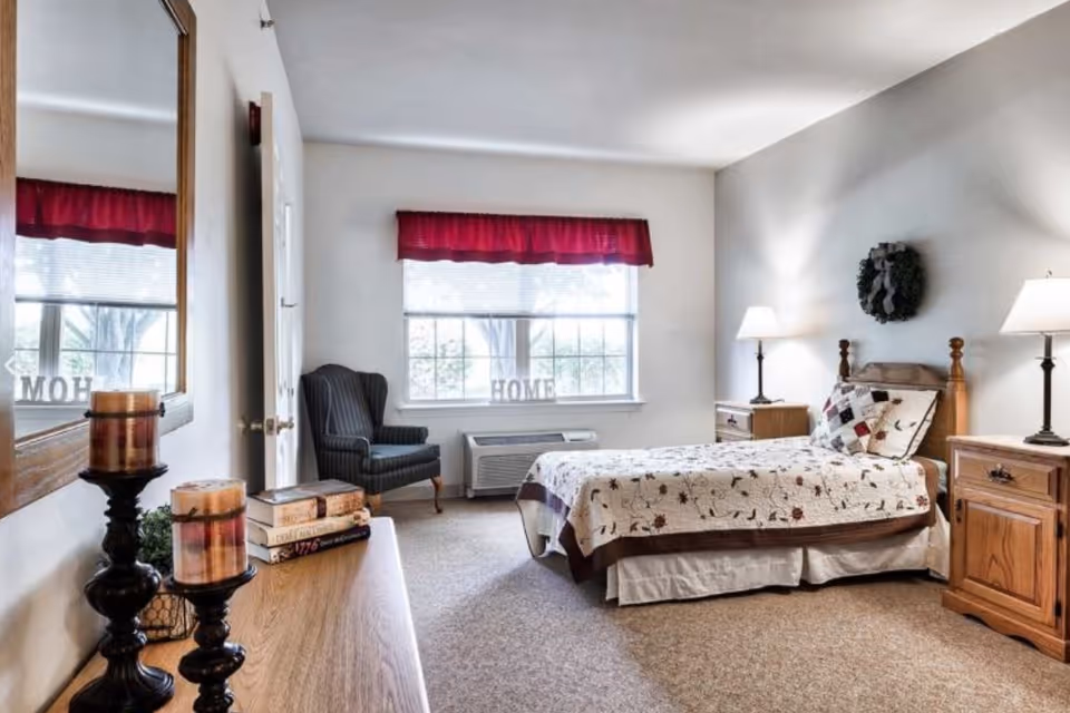 A cozy bedroom with a single bed covered in a floral quilt, two wooden nightstands each with a lamp, a dark upholstered armchair near a window with a red valance, and a wooden dresser with candles and books. A wreath hangs on the wall above the bed.
