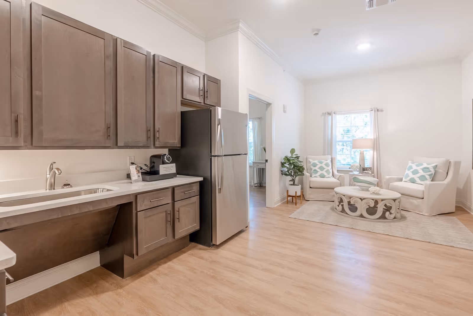 A bright and clean interior space featuring a kitchenette with wooden cabinets, a stainless steel refrigerator, and a sink on the left side. On the right side, there is a small sitting area with two white armchairs with patterned cushions, a round decorative coffee table, a floor lamp, and a window with light curtains. The floor is light wood, and the walls are painted white.