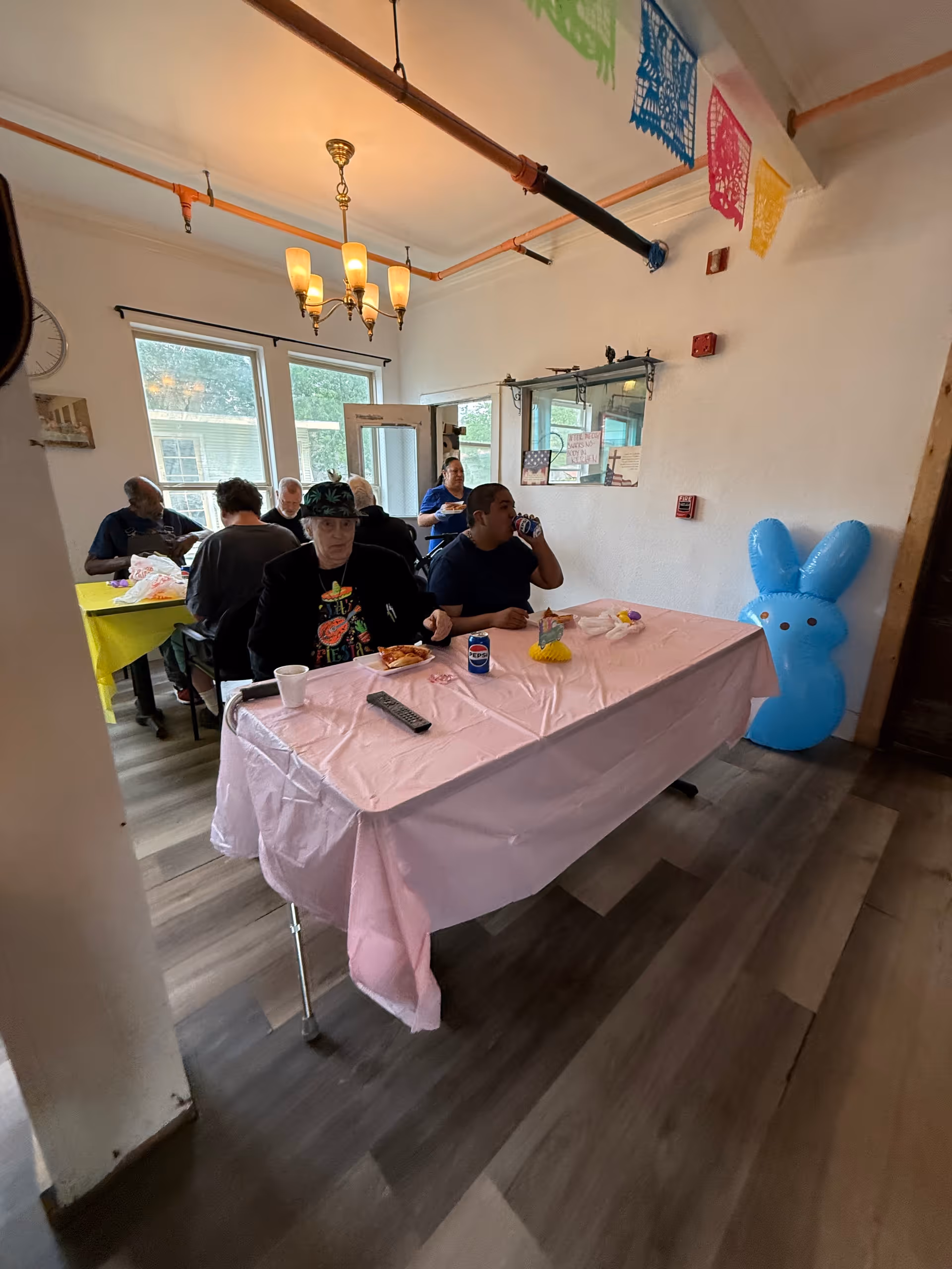 Residents seated at decorated tables eating and drinking in a communal dining room with hanging lights and festive decorations.