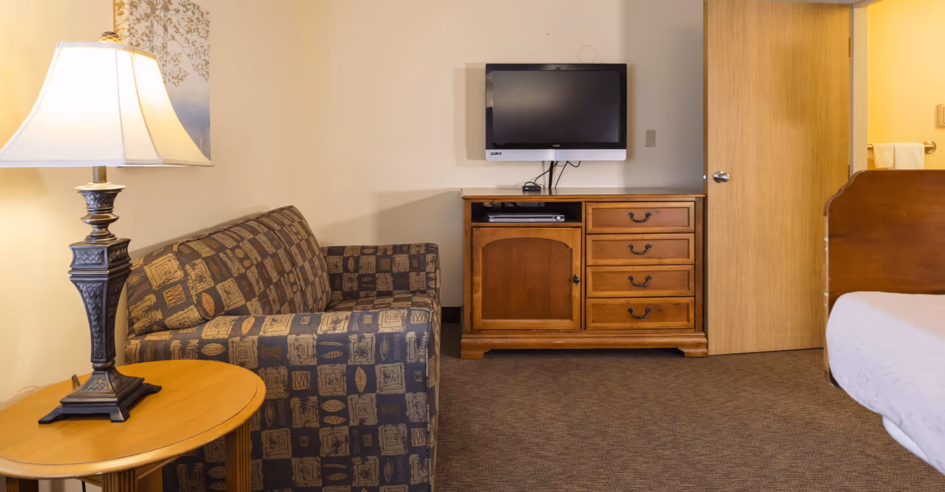 A cozy room with a patterned armchair next to a wooden side table with a decorative lamp. Across from the chair is a wooden dresser with drawers and a cabinet, topped with a flat-screen TV mounted on the wall. A partially visible bed with white bedding is on the right side, and a wooden door is in the background.