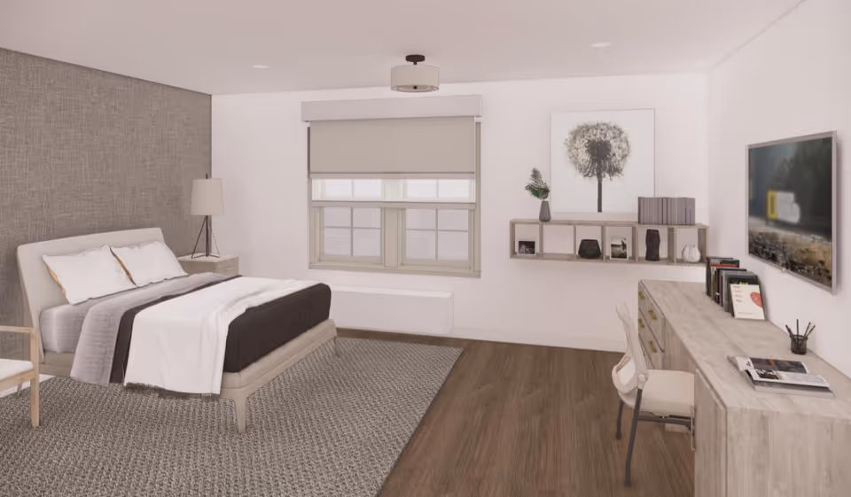 A modern and cozy bedroom in an assisted living facility featuring a bed with white and gray bedding, a bedside table with a lamp, a large window with a roller shade, a wall-mounted shelf with decorative items and books, a desk with a chair, books, and a flat-screen TV mounted on the wall. The room has wood flooring and a textured gray accent wall behind the bed.