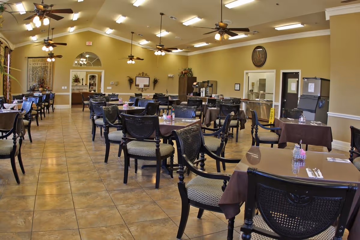A spacious dining room with multiple tables covered in brown tablecloths, each set with utensils, napkins, and condiments. The room has beige walls, tiled floors, ceiling fans with lights, and large windows with curtains. There is a clock on the wall and a serving area with coffee machines and other equipment in the background.