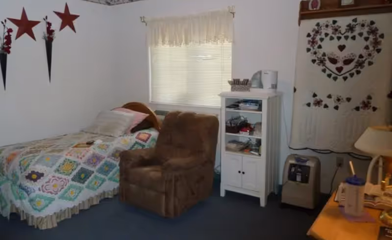Small bedroom with a single bed covered by a patchwork quilt, a brown recliner, a white shelving cabinet, and decorative wall hangings.