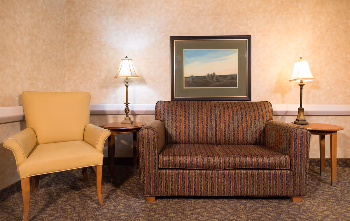 A cozy sitting area with a patterned loveseat and a yellow armchair separated by two wooden side tables, each holding a lit table lamp. A framed landscape painting hangs on the beige textured wall behind the loveseat.