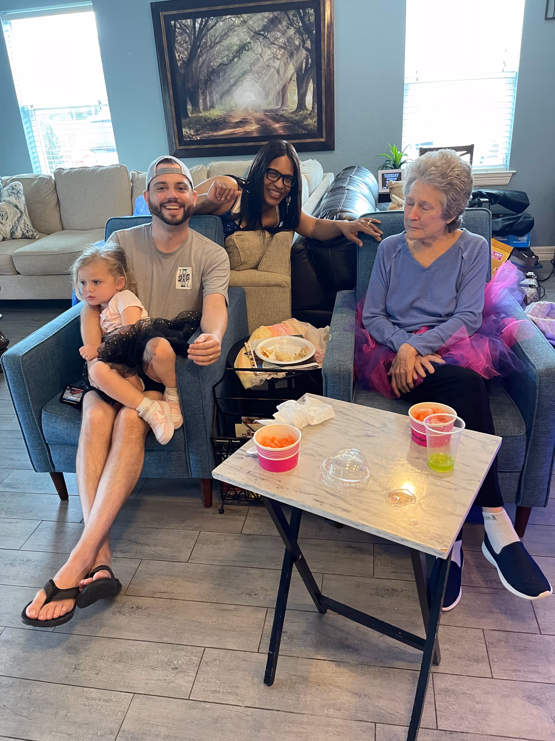 A group of four people sitting in a living room area. A man wearing a cap and flip-flops is sitting on a blue armchair holding a young girl dressed in a pink top and black tutu. Next to them, a woman with glasses is leaning on the armchair smiling. An elderly woman wearing a purple top and a pink tutu is sitting on another blue armchair. In front of them is a small table with cups containing snacks and drinks. The background shows a beige sofa, a framed picture of a tree-lined path, and windows letting in natural light.