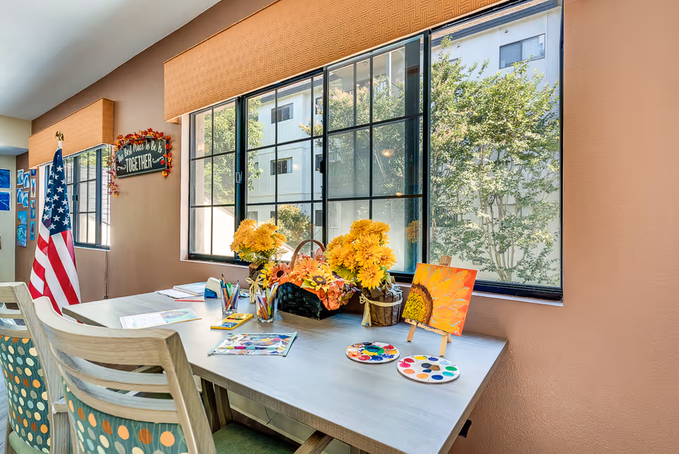 A bright room with a large window showing trees outside. A table is set up with art supplies including paint palettes, colored pencils, and a small easel holding a painting of a sunflower. There are two chairs with patterned cushions, a basket with yellow flowers, and an American flag standing in the corner. A wall decoration reads 'The best place to be is TOGETHER'.