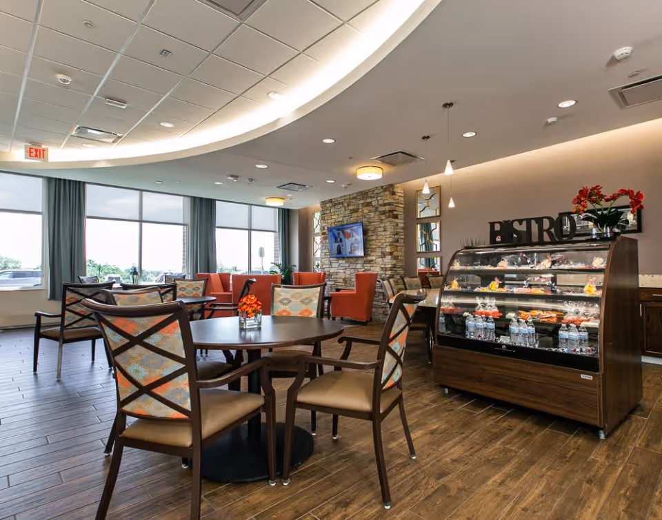 Spacious bright dining and lounge area with round tables and chairs, a pastry display case labeled 'BISTRO', and seating by large windows.