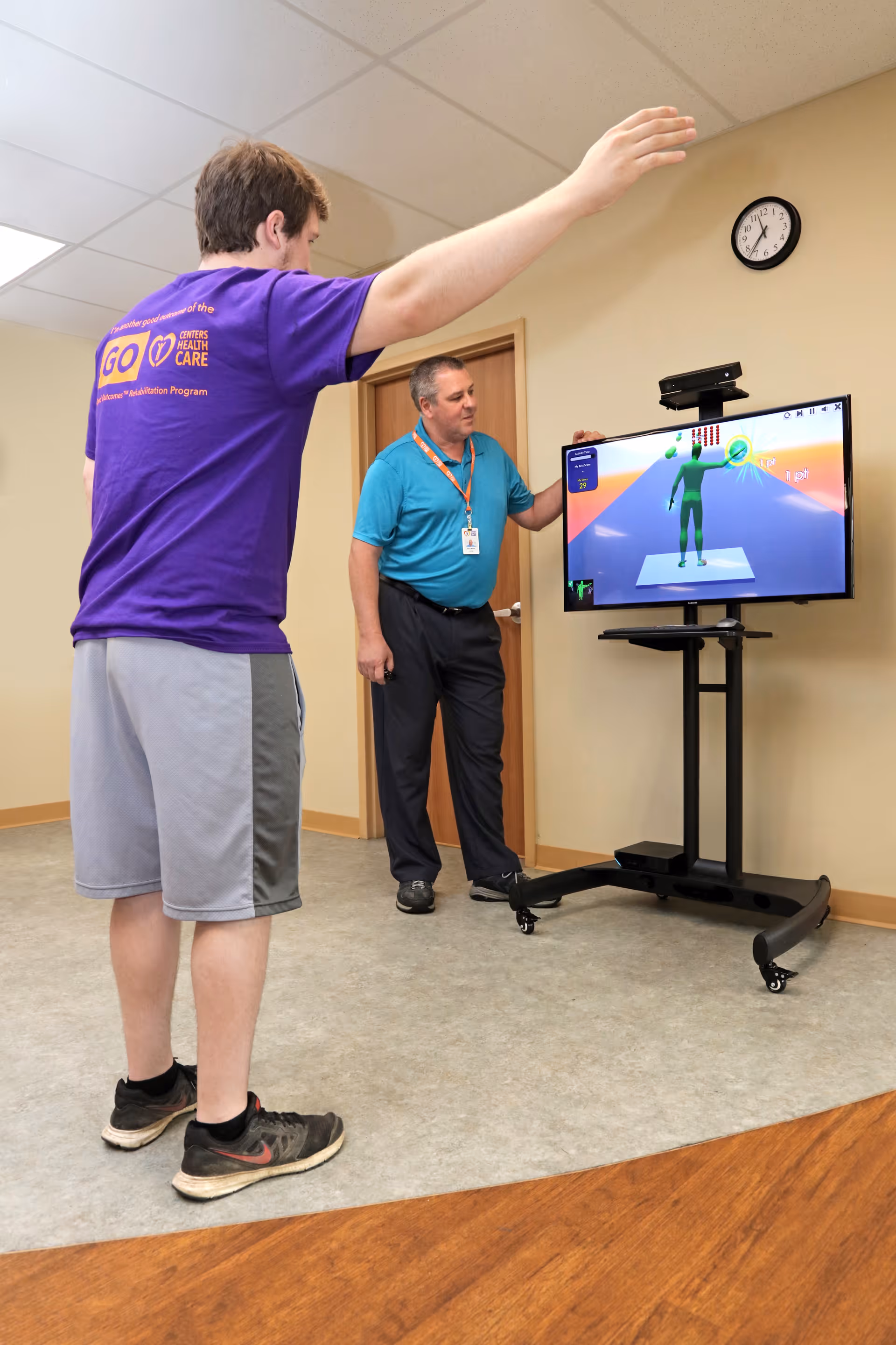 A man in a purple shirt and gray shorts is standing with one arm raised, facing a screen displaying a virtual figure. Another man in a blue shirt and black pants stands nearby, observing the screen. The setting appears to be an indoor room with beige walls and a clock on the wall.