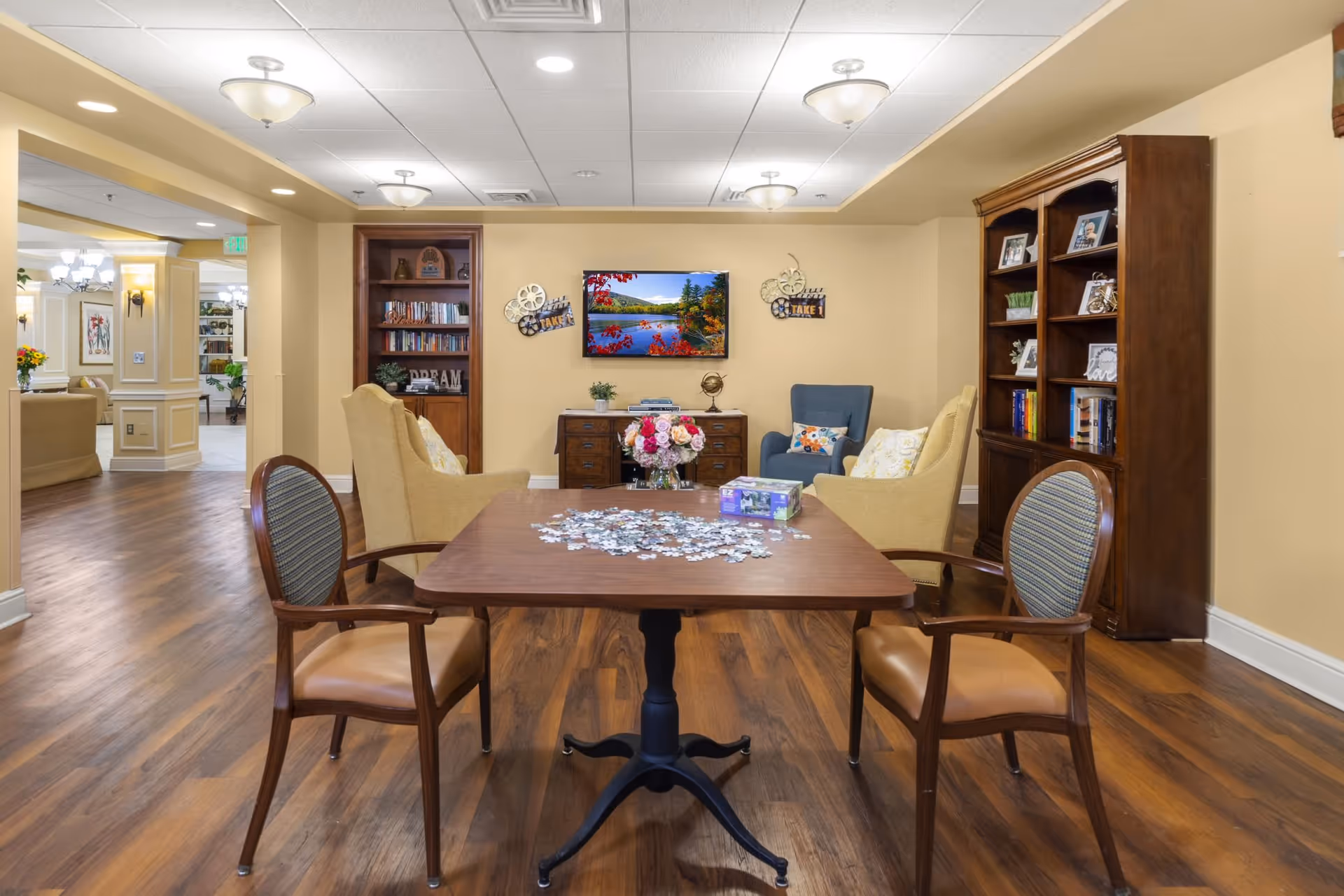 A cozy common area in a senior living facility with a wooden table in the center holding a partially completed jigsaw puzzle and a box. Surrounding the table are four chairs with cushioned seats and backs. In the background, there are two beige armchairs and one blue armchair arranged around a wooden cabinet with a flat-screen TV mounted above it. The walls are painted light yellow, and there are two wooden bookshelves filled with books and decorative items. The floor is wood, and the ceiling has recessed lighting and ceiling fixtures.