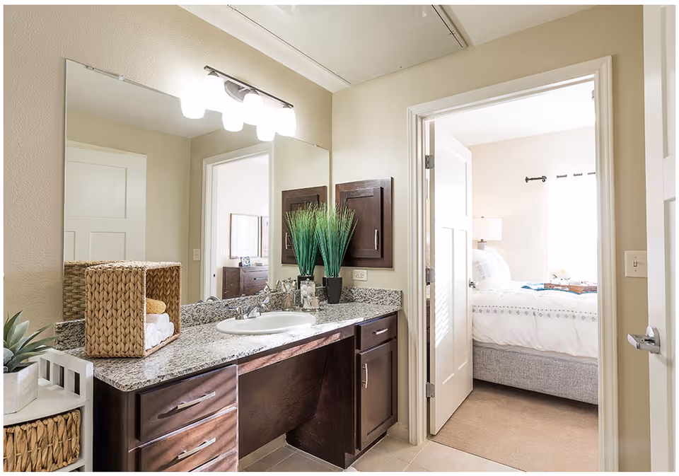 A bathroom vanity area with a granite countertop, a sink, and a large mirror above. There are decorative plants and a woven basket with towels on the counter. The bathroom opens into a bedroom with a neatly made bed, a bedside lamp, and a window with curtains.