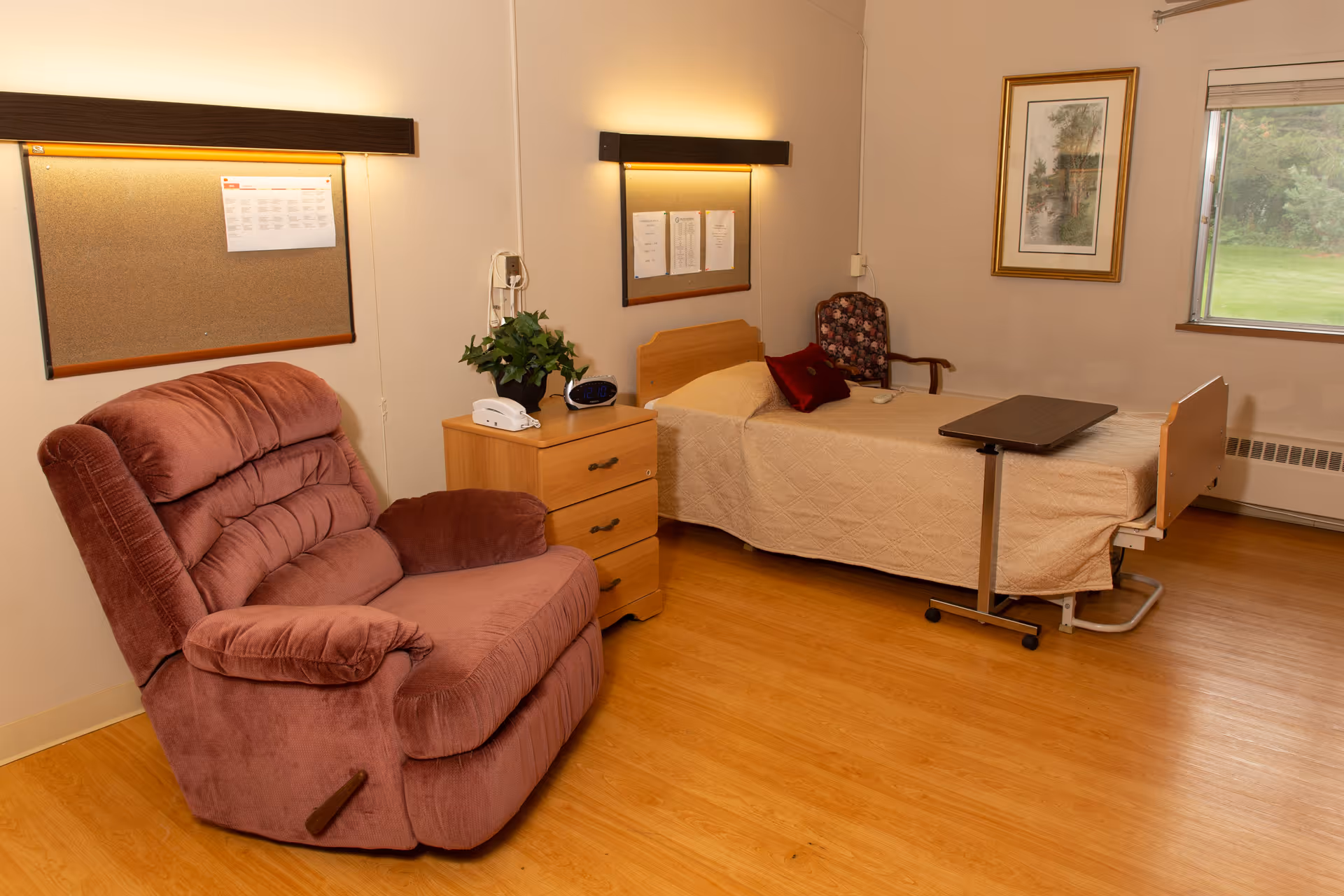 A senior living facility room with a single bed covered in a beige quilt, a brown reclining chair, a wooden nightstand with a telephone and a plant, a floral armchair, a framed picture on the wall, two bulletin boards with papers pinned, and a window showing greenery outside.