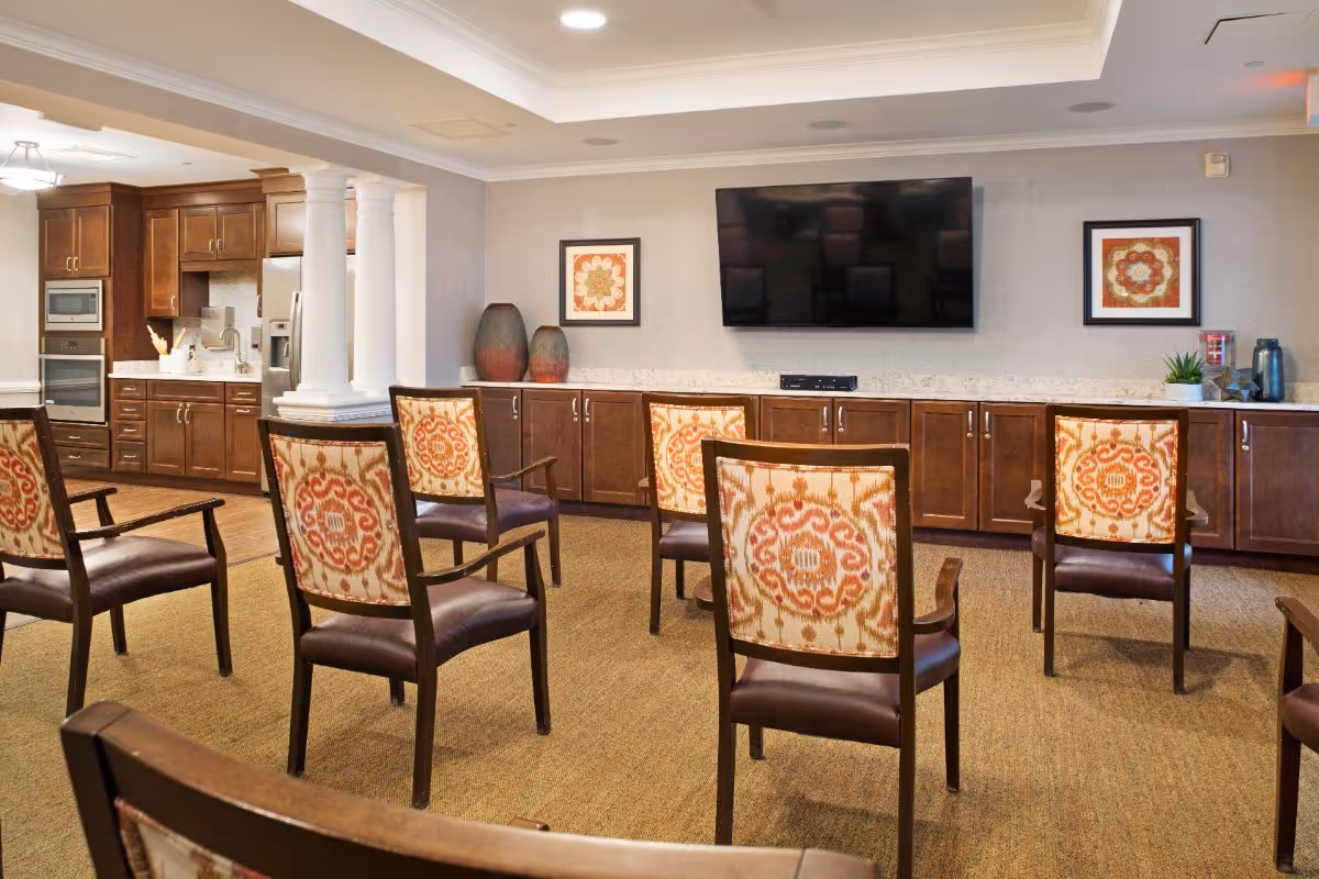 A senior living common room with patterned chairs arranged facing a wall-mounted TV and cabinets with a kitchenette in the background.