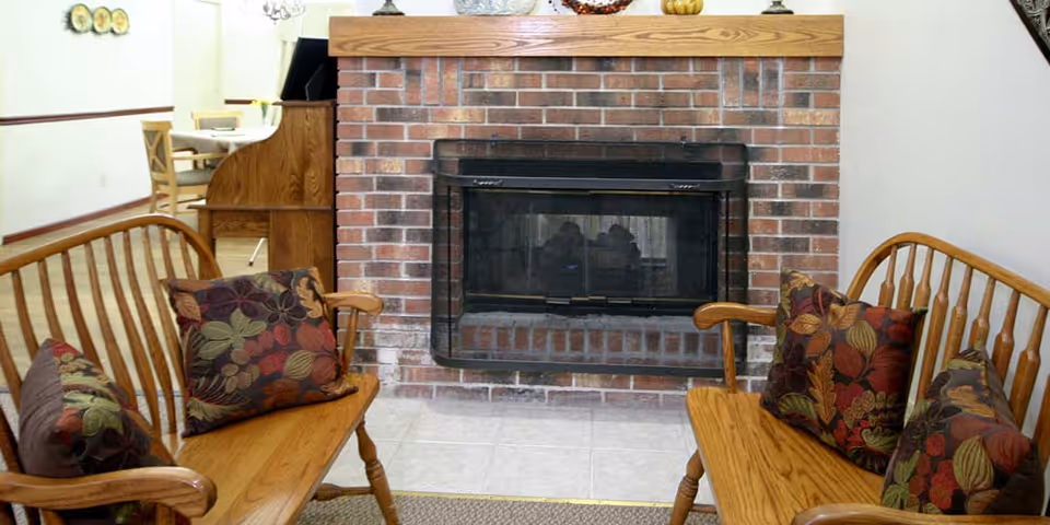 A cozy interior room featuring a brick fireplace with a black screen, flanked by two wooden benches with floral patterned cushions. In the background, there is a wooden desk and chairs, and a wall with decorative plates.