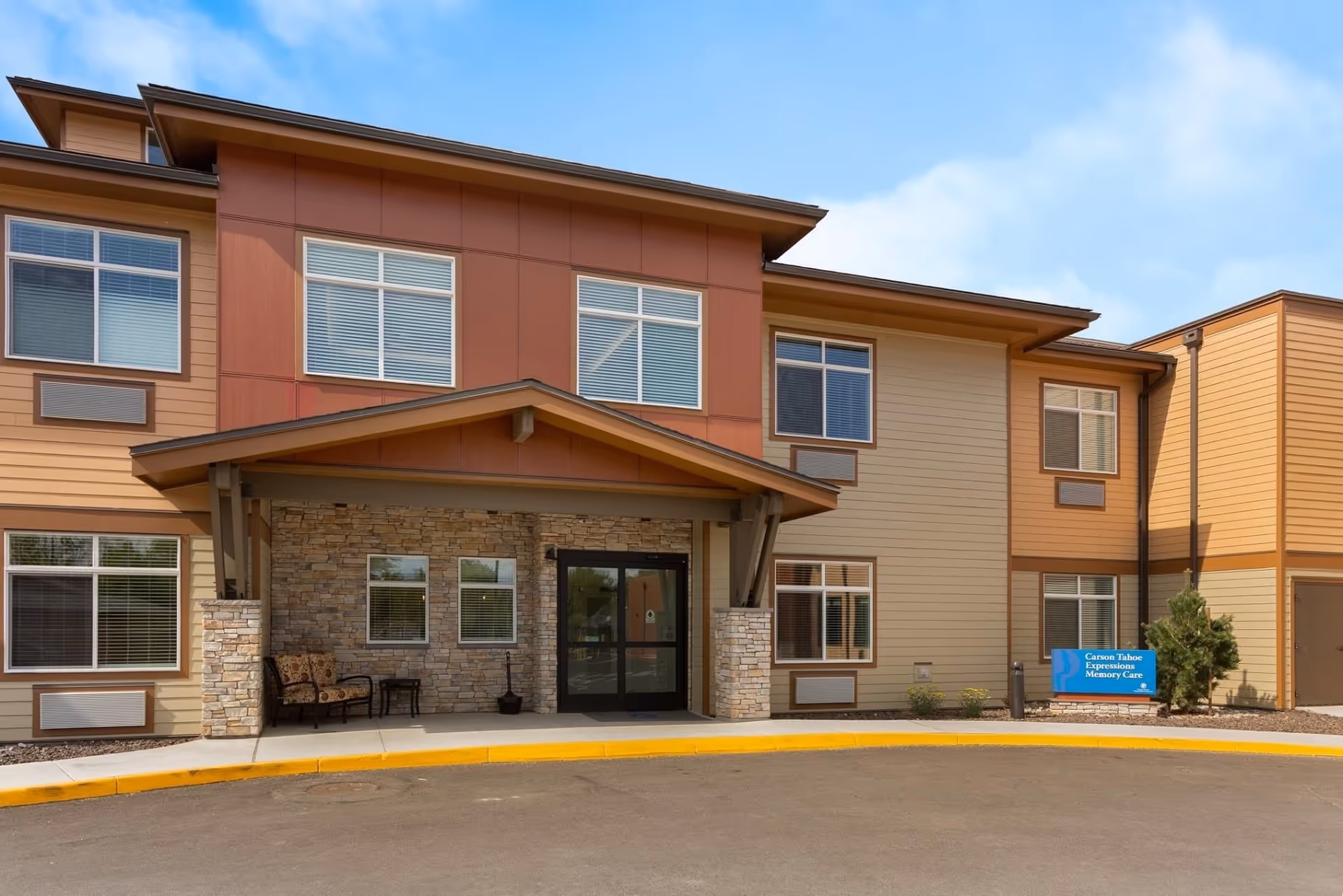 Exterior front view of a two-story senior living facility building with beige and brown siding, stone accents around the entrance, large windows, and a covered entryway with two chairs and a small table on the porch. A blue sign near the entrance reads 'Carson Tahoe Expressions Memory Care'.