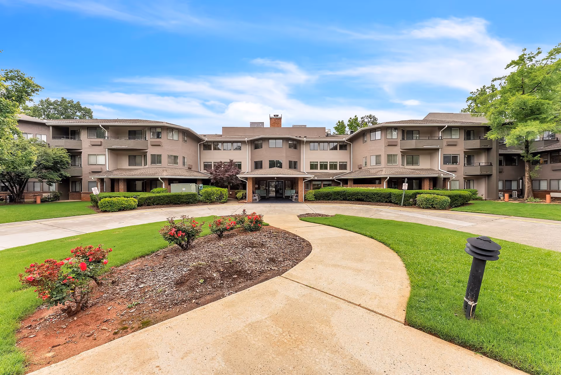 Front exterior view of Regency House by Barclay House, a multi-story senior living facility with a curved driveway, landscaped garden with red flowers, green lawns, and trees under a blue sky with some clouds.