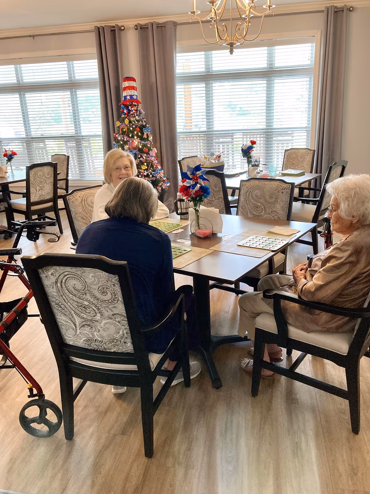 Three elderly women sitting around a table in a well-lit room with large windows and gray curtains. The table has placemats, a vase with red, white, and blue flowers, and bingo cards. A decorated Christmas tree with a patriotic theme stands near the window. One woman has a walker next to her chair.