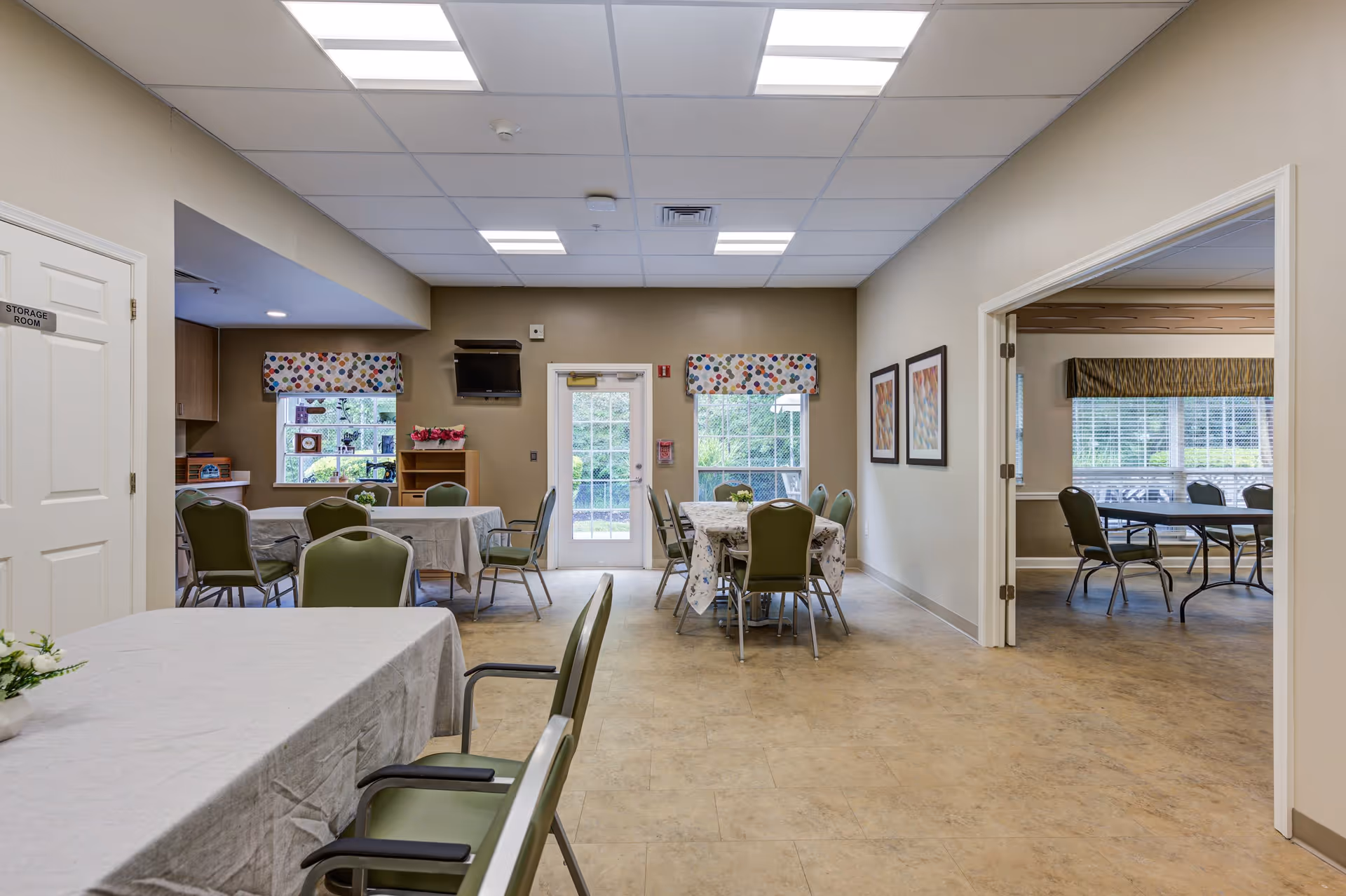 Interior view of a dining area in a senior living facility with several tables covered with tablecloths and surrounded by green chairs. There are windows with colorful valances, a door leading outside, framed artwork on the walls, and a small wall-mounted TV. Adjacent room visible with more tables and chairs.
