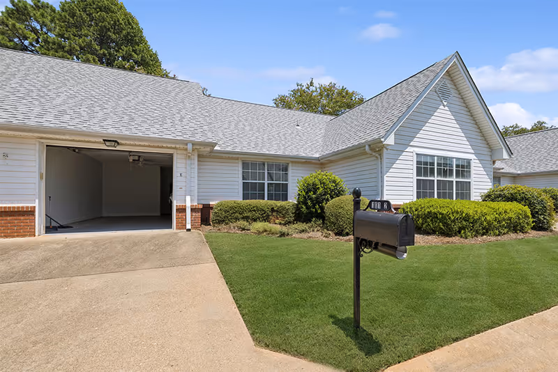 Exterior view of a single-story residential building with white siding and a gray shingled roof. There is an open garage on the left side, a well-maintained green lawn, bushes, and a black mailbox in the foreground. The sky is clear with a few clouds.
