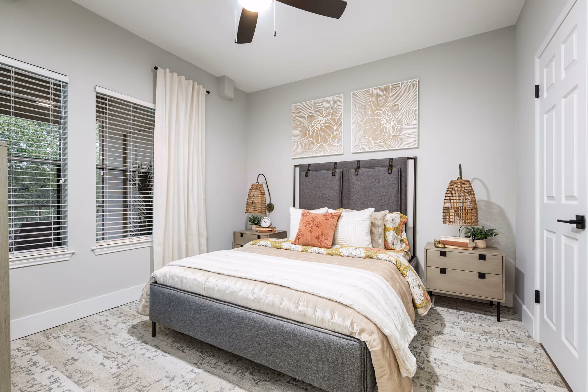 Well-lit bedroom with a upholstered bed, patterned bedding and pillows, two nightstands with wicker lamps, wall art above the headboard, and windows with blinds.