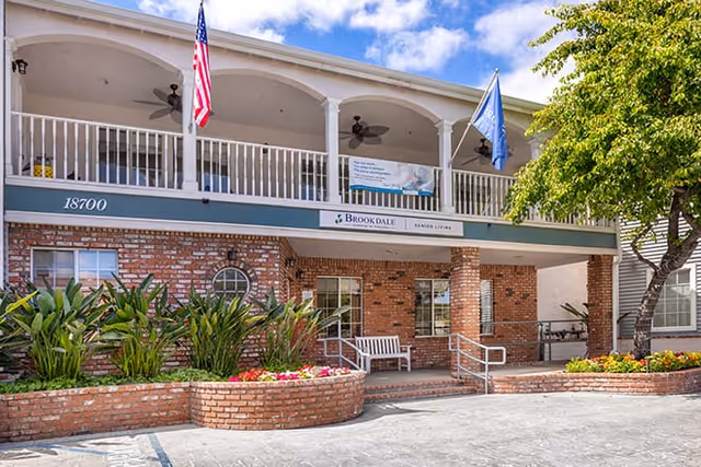 Exterior view of Brookdale Gardens of Tarzana senior living facility showing a two-story brick building with a balcony, American and facility flags, landscaped flower beds, and a bench near the entrance.