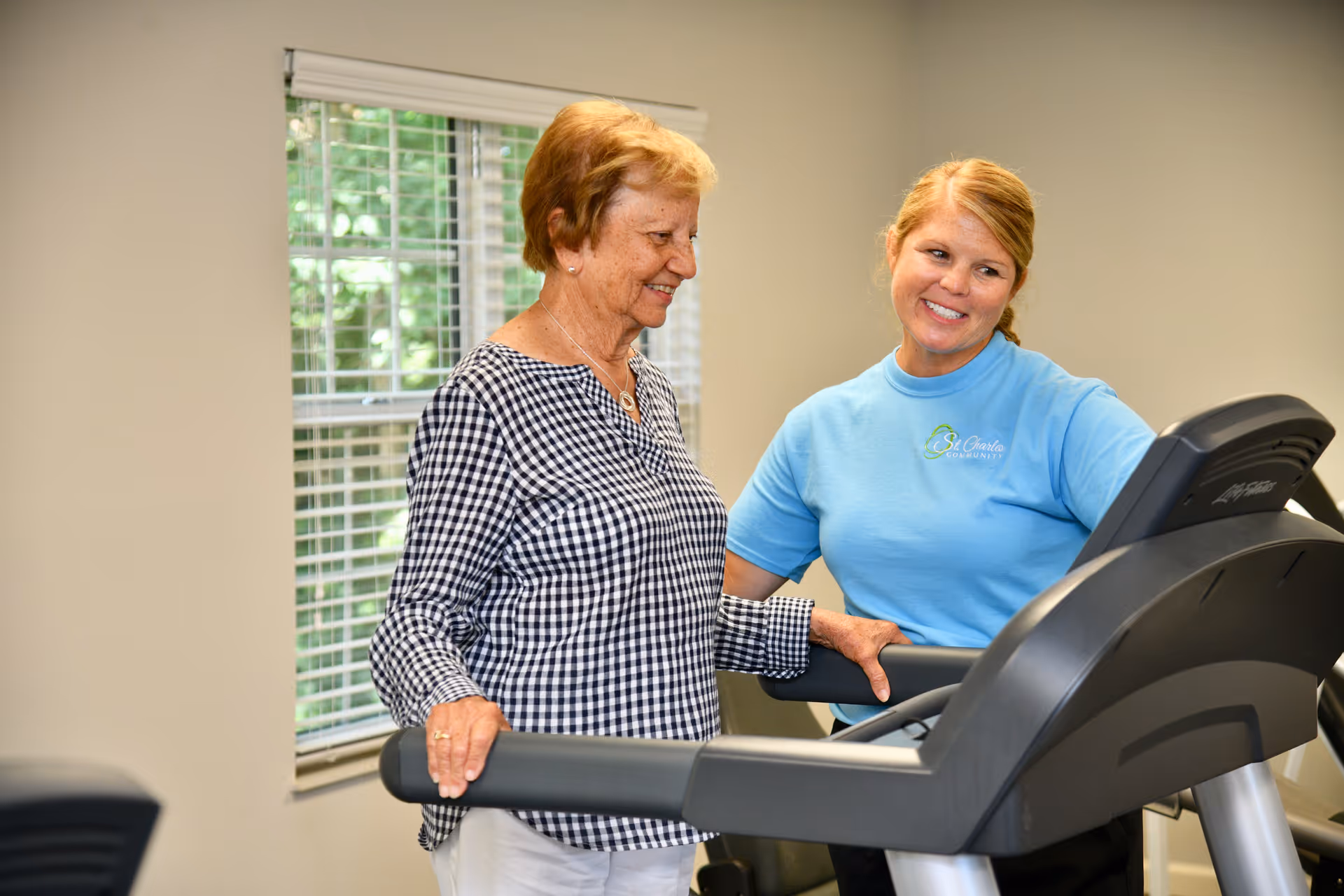 An elderly woman using a treadmill in a fitness room with a staff member from St. Charles Community smiling and assisting her.
