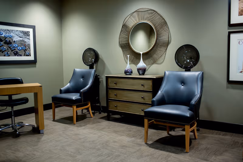 A modern interior room with two dark blue leather chairs with wooden legs, a wooden dresser with three drawers, decorative vases on top, and a round mirror with a unique frame hanging above the dresser. There is a wooden desk with a black office chair on the left side and framed artwork on the walls.