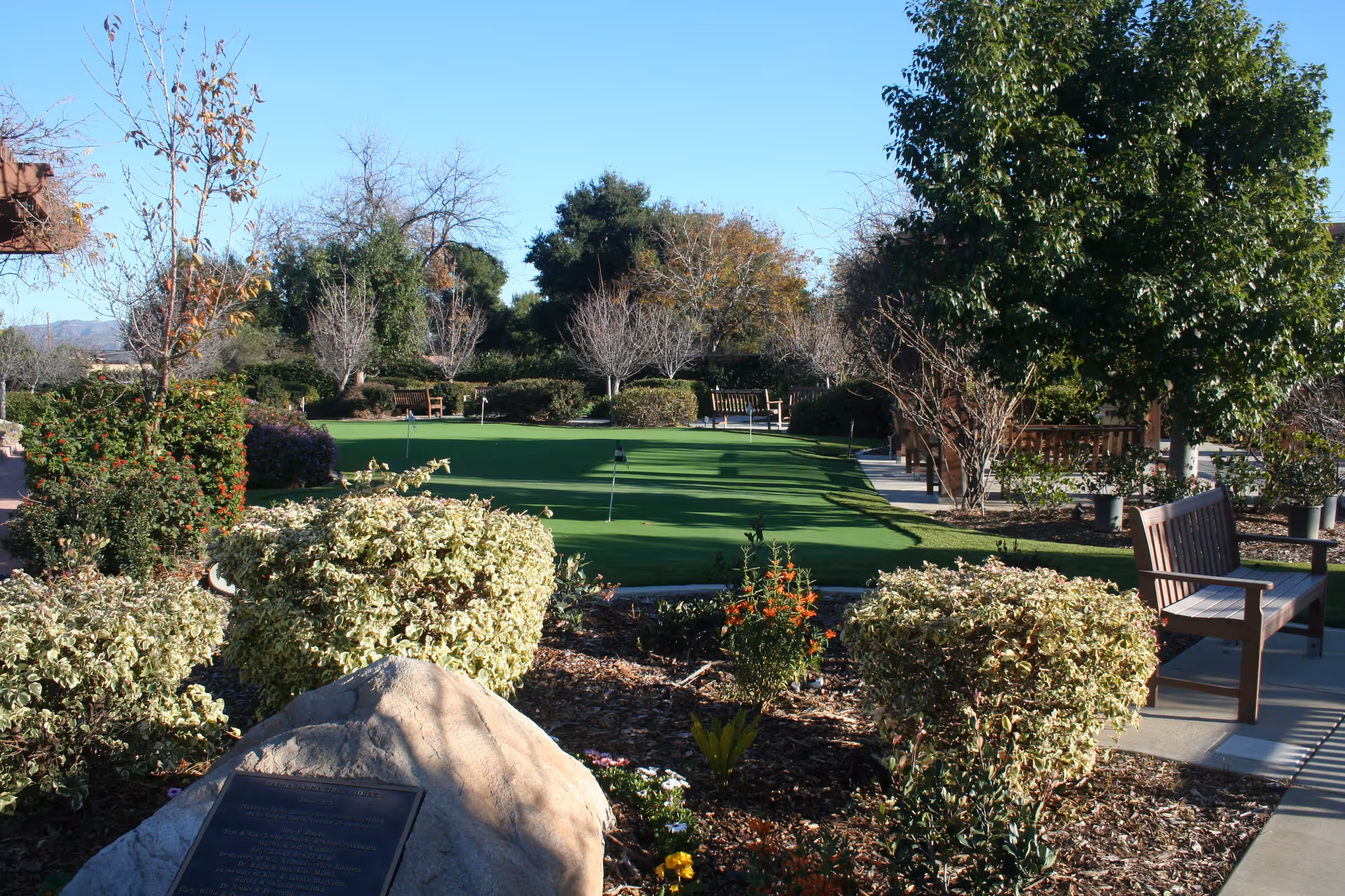 A well-maintained outdoor garden area with a putting green surrounded by various bushes, trees, and flower beds. Several wooden benches are placed along the paved pathways, and a large rock with a commemorative plaque is visible in the foreground. The sky is clear and blue.