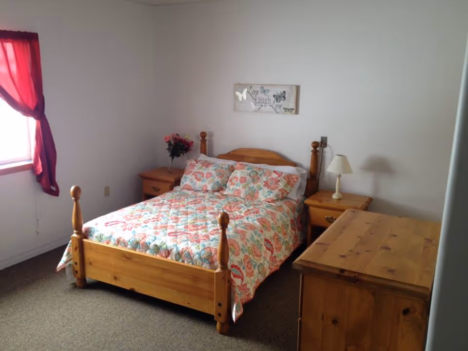 A simple bedroom with a wooden bed dressed in a floral quilt, two matching nightstands, a dresser, and a window with red curtains.