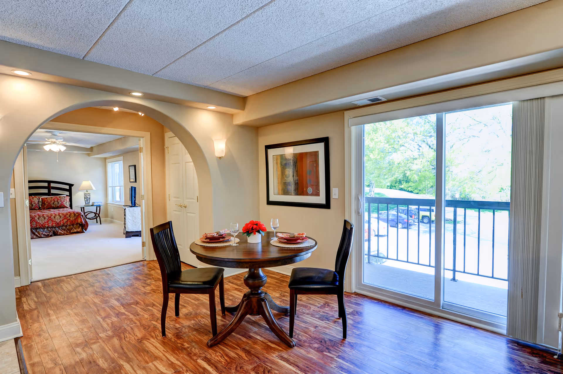 A cozy dining area with a round wooden table set for two with plates, glasses, and a small flower centerpiece. The room has wooden flooring and a large sliding glass door leading to a balcony with a view of trees and parked cars. Through an arched doorway, a bedroom with a bed, nightstand, lamp, and ceiling fan is visible.