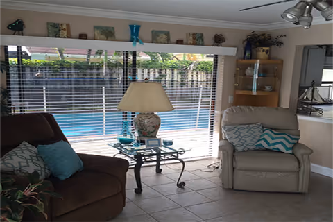 A cozy living room with a brown armchair and a beige recliner, both adorned with patterned cushions. Between them is a glass-top side table holding a decorative lamp and small items. Large sliding glass doors with blinds reveal a swimming pool and greenery outside. Shelves with decorative items are visible on the right side of the room.