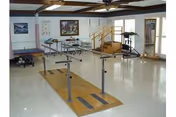 Bright rehabilitation room featuring parallel walking bars, a treadmill, stairs, and other therapy equipment.