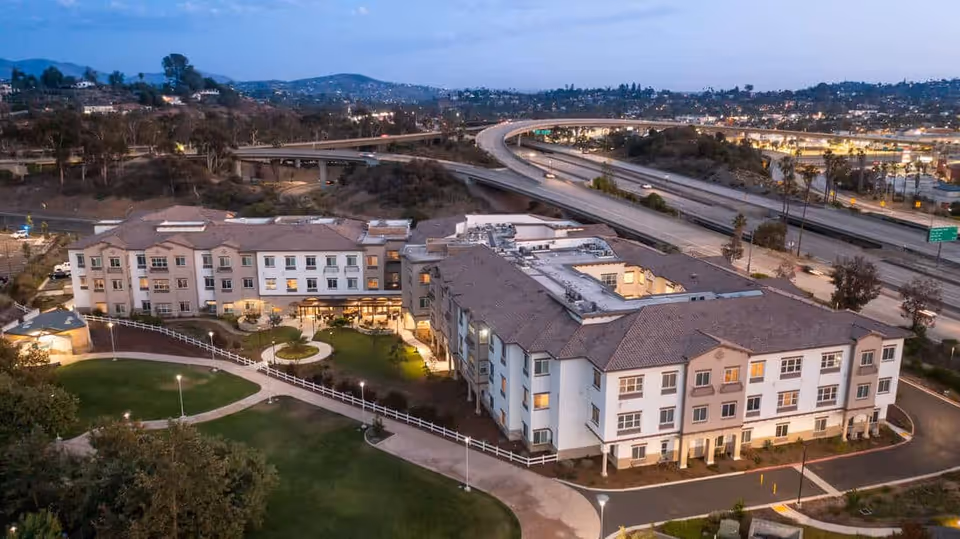 Aerial view of a three-story senior living facility with landscaped grounds beside a freeway at dusk.