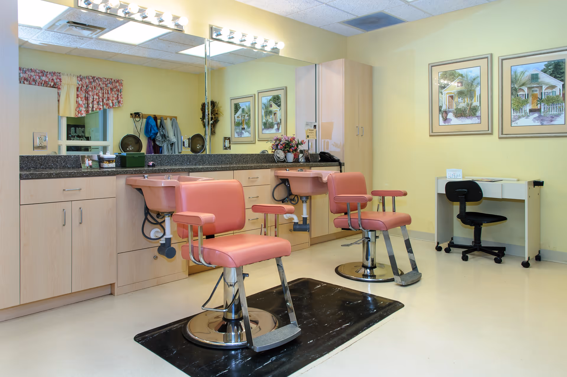 Interior view of a salon area in a senior living facility with two pink salon chairs in front of sinks and a large mirror. The room has light yellow walls, a countertop with cabinets underneath, framed pictures on the wall, and a small desk with a black office chair.
