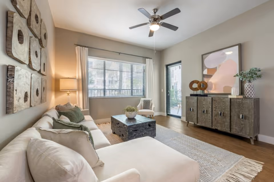 A bright and modern living room with a large beige sectional sofa adorned with green and beige pillows. A wooden coffee table sits on a textured area rug in front of the sofa. On the wall above the sofa are six square wooden art pieces with circular designs. A floor lamp with a beige shade is positioned in the corner near a large window with white curtains and blinds. Across from the sofa is a wooden sideboard decorated with abstract art, two circular wooden sculptures, a small plant in a patterned vase, and a white decorative object. A ceiling fan with lights is mounted on the white ceiling, and a glass door leads to an outdoor area.