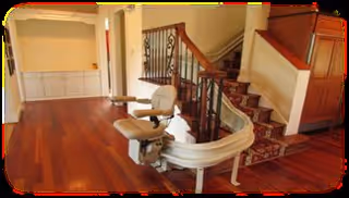 Interior view of a senior living facility featuring a wooden staircase with a stair lift installed. The area has polished hardwood floors, a built-in cabinet with shelving on the left, and warm beige walls. The stair lift is positioned at the base of the stairs, which are carpeted with a patterned runner.