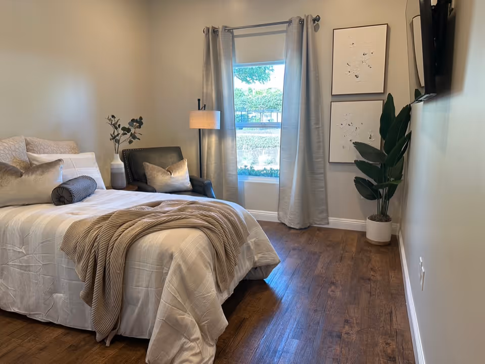 A cozy bedroom with a neatly made bed featuring white and beige bedding, a beige throw blanket, and multiple pillows. Next to the bed is a black armchair with a beige pillow and a floor lamp. A window with gray curtains lets in natural light, and two framed floral artworks hang on the wall beside a potted plant. The room has wooden flooring and a wall-mounted TV.
