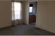 Empty carpeted living area with a window with blinds and curtains and a doorway leading to a sink/bathroom area.