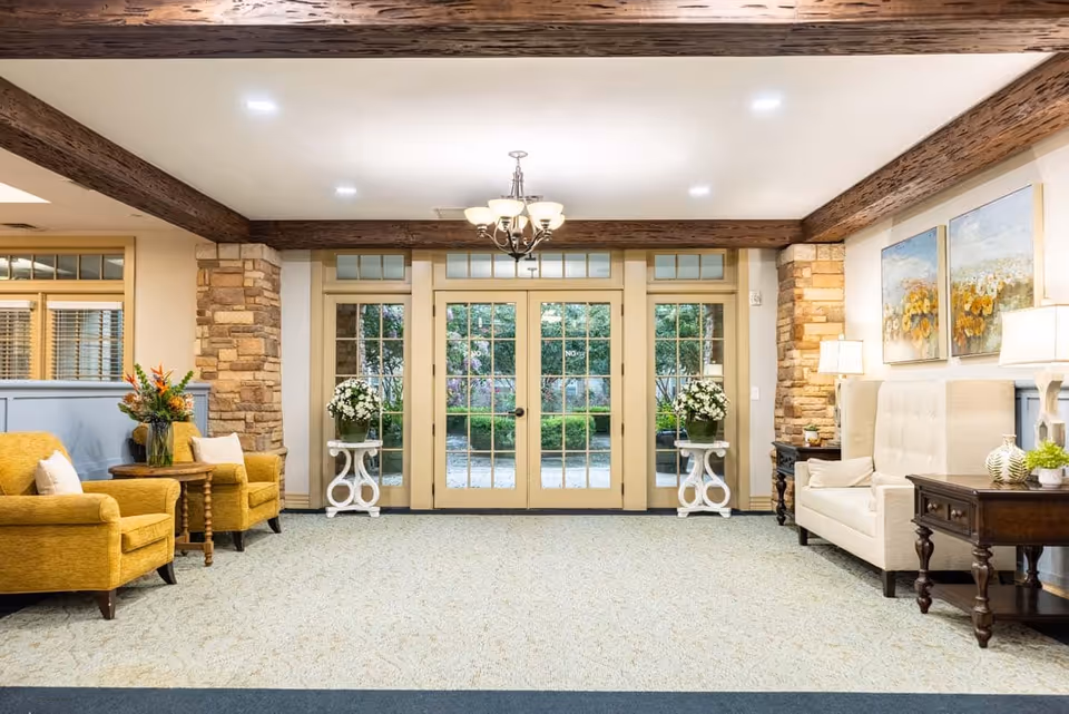 Well-lit lounge with yellow armchairs and a beige sofa flanking glass double doors, exposed wooden beams, stone columns, and side tables with flowers.