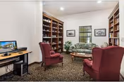 A cozy interior room with two red upholstered armchairs, a patterned sofa, wooden bookshelves filled with books, a small wooden coffee table, a computer desk with a monitor, and framed pictures on the walls.