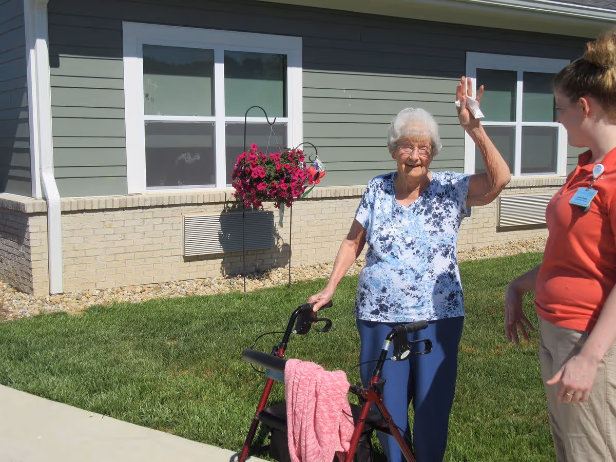 An elderly woman standing outside on a grassy area next to a building, holding a walker with a pink cloth draped over it and raising one hand. A younger woman wearing a red shirt and name badge stands nearby, smiling at her. There is a hanging basket of pink flowers attached to the building wall behind them.