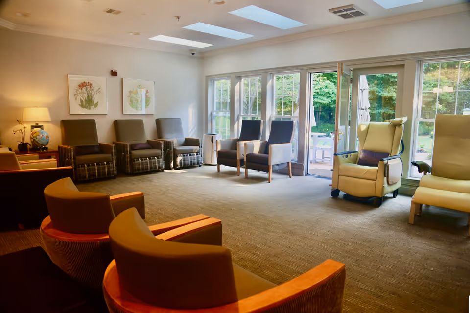 A bright memory-care common room with multiple armchairs arranged facing large windows and glass doors opening to a garden.