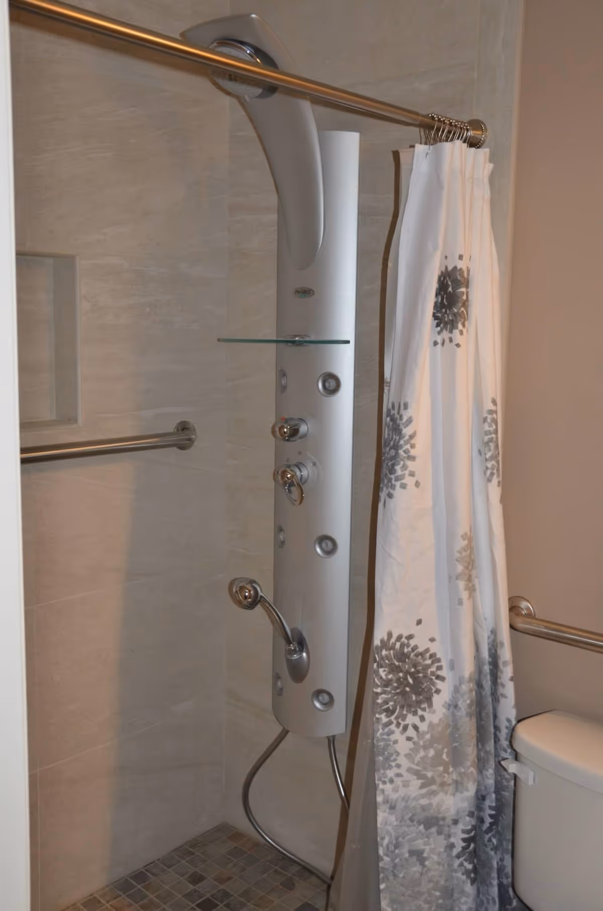 A bathroom shower area with a modern vertical shower panel featuring multiple controls and jets, a handheld showerhead, a glass shelf, and a metal grab bar on the tiled wall. A white shower curtain with gray floral patterns is partially drawn, and a toilet with a metal grab bar beside it is visible.