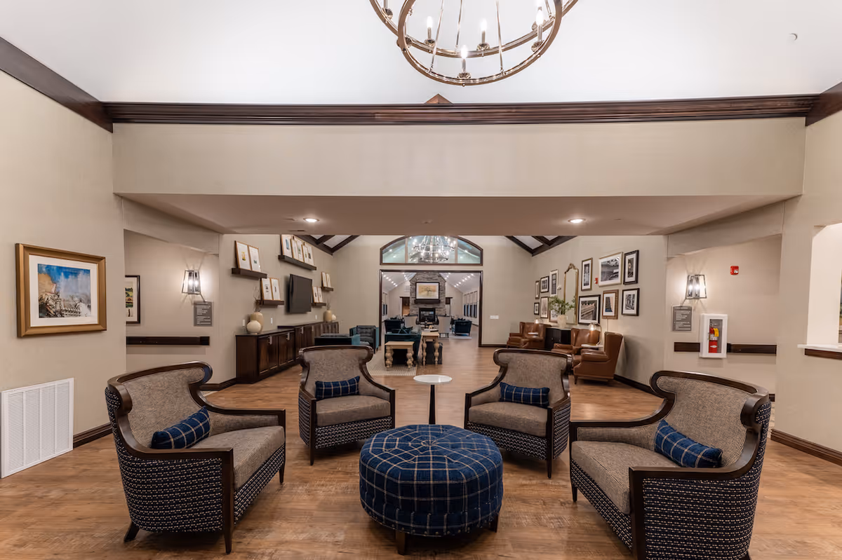 A spacious and well-lit senior living facility common area with four cushioned armchairs arranged around a round blue plaid ottoman. The room features wooden flooring, beige walls with framed artwork, and a chandelier hanging from the ceiling. In the background, there is a hallway leading to another seating area with additional chairs and a fireplace.